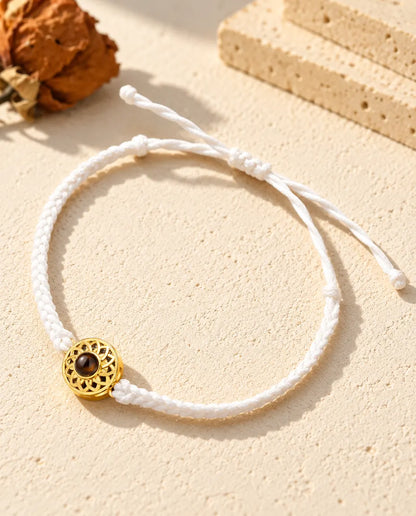 Armband mit schmalem weissem Flechtband, goldfarbenem Rund-Charm und Schiebeknoten auf hellem Gips