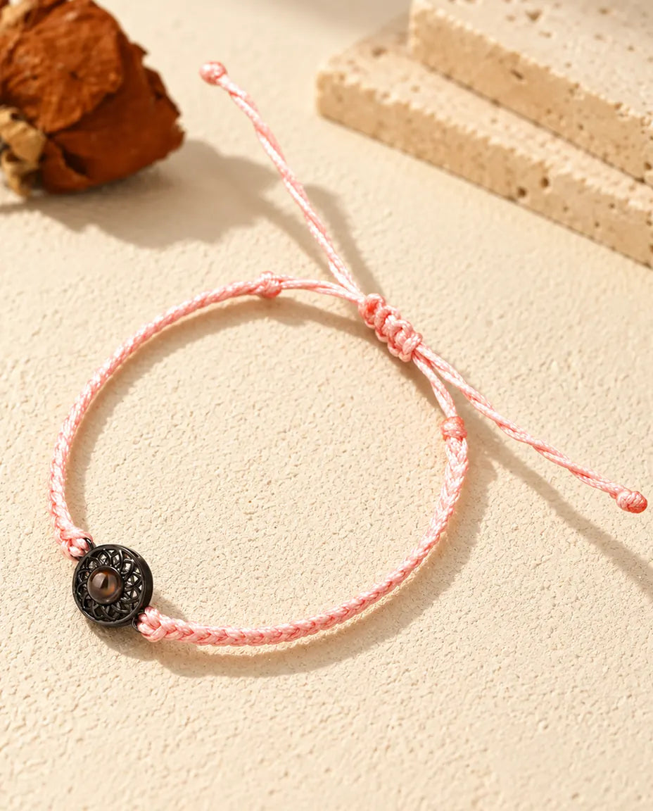 Armband mit schmalem rosa Flechtband, schwarzem Rund-Charm und braunem Bildstein auf hellem Gips
