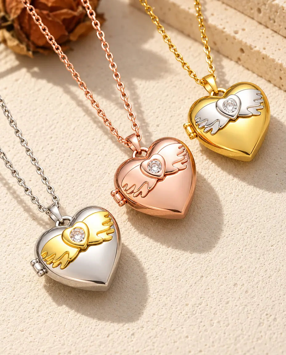 Herz-Foto-Medaillon mit Engelsflügeln in Silber-, Roségold- und Goldfarbe schräg auf cremefarbenem Putz